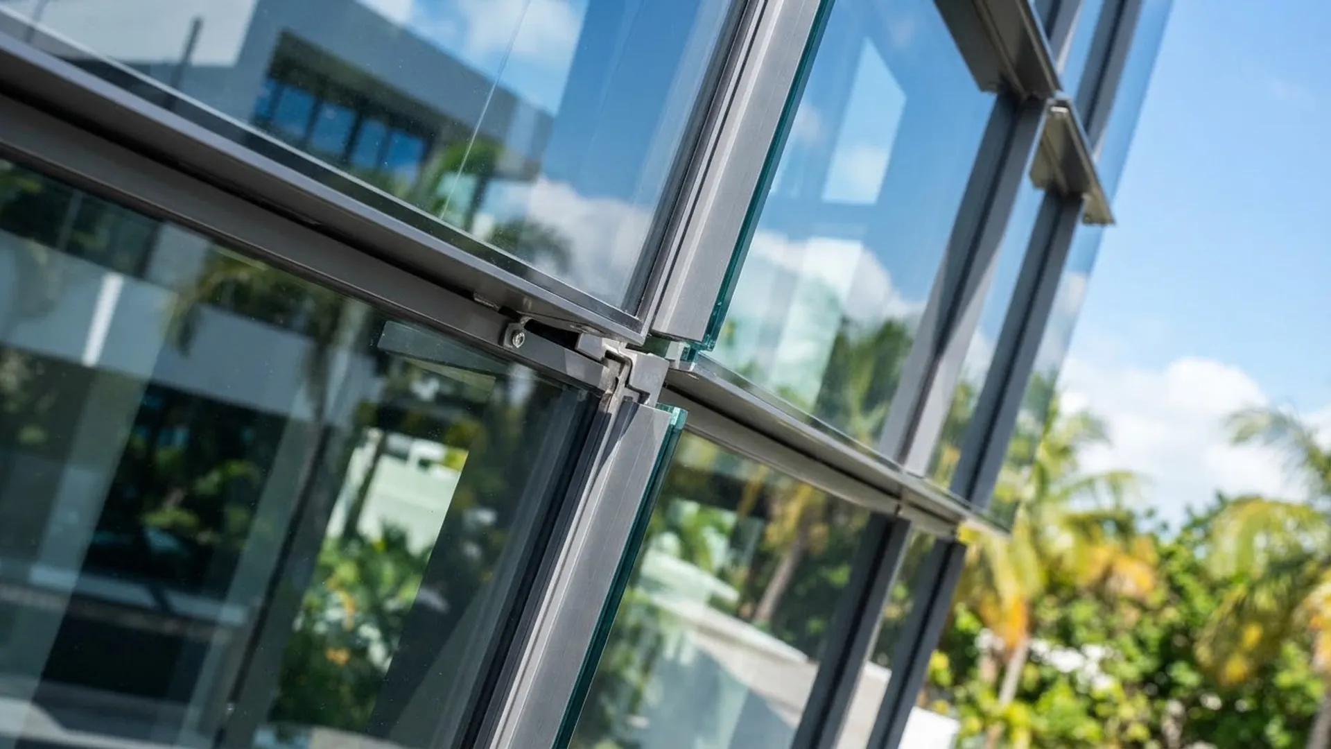 Glazed façade corner carried on aluminium framing, with structural fittings visible against tropical planting.